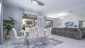Una sala de estar moderna con un sofá gris, una mesa de comedor de vidrio y sillas blancas, y un televisor grande en la pared.