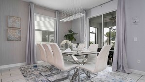Un comedor moderno con una mesa de vidrio y sillas blancas, una araña y vistas a través de amplios ventanales.