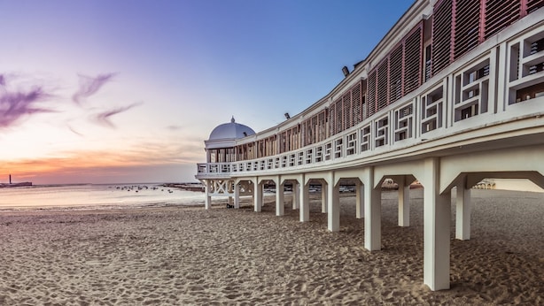 Un muelle frente a la playa con una estructura blanca y un edificio de forma abovedada al fondo.