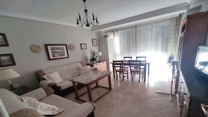 Una sala de estar con un sofá, una mesa de centro y un comedor con sillas y una mesa. Hay un televisor a la derecha.