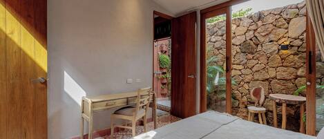 A bedroom with a bed, a desk, and a chair. There is a stone wall and a door leading to an outdoor area.
