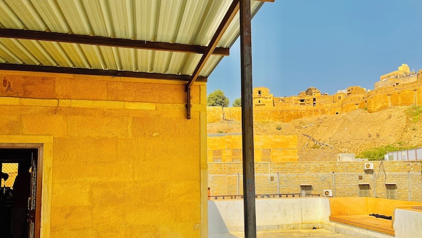 A building with a yellow exterior, a metal roof, and a view of a hill with ancient structures. - Hotel Royal Queen Jaisalmer  (Jaisalmer)