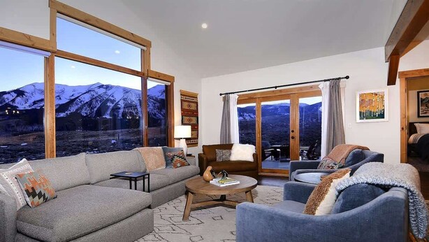Ein Wohnzimmer mit einer Couch, Sesseln, einem Couchtisch und großen Fenstern mit Blick auf die Berge.