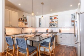 A modern kitchen with white cabinetry, a central island with chairs, pendant lighting, and a stainless steel refrigerator.