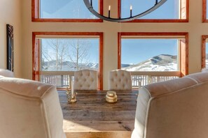 A dining area with a wooden table, two upholstered chairs, a chandelier, and large windows with a mountain view.