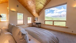Ein Schlafzimmer mit einem großen Bett, zwei Fenstern, einem Schreibtisch und Blick auf eine gebirgige Landschaft.