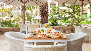 An outdoor dining area with wicker chairs and tables, surrounded by palm trees and a pool.