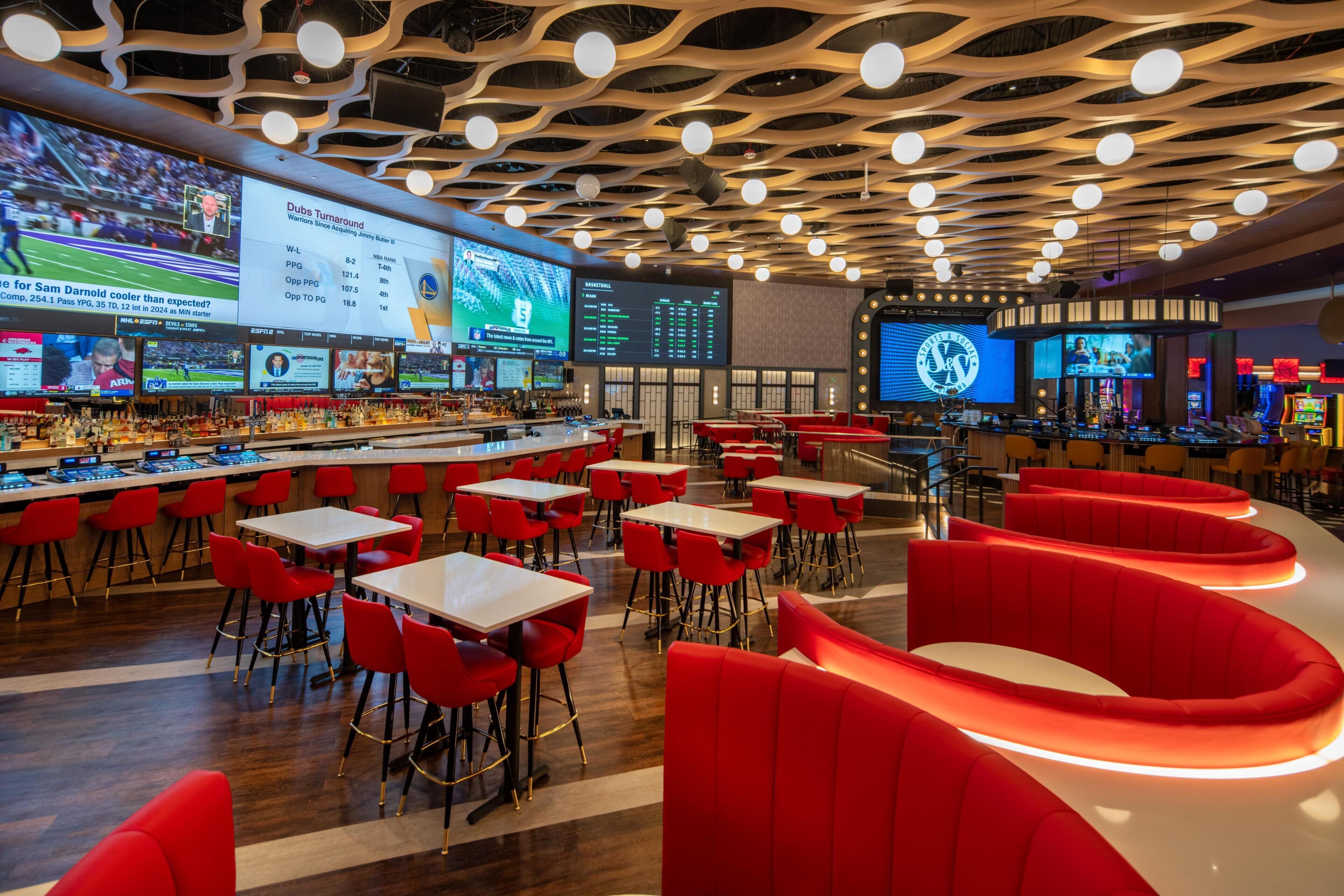 A modern bar area with multiple screens displaying sports content, red seating, and a unique ceiling design.