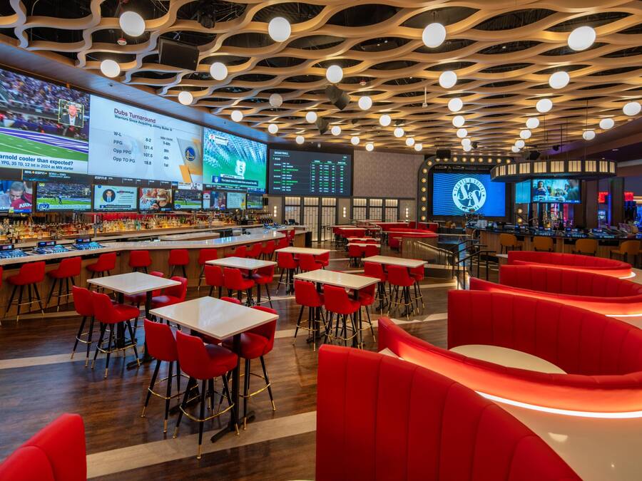 A modern bar area with multiple screens displaying sports content, red seating, and a unique ceiling design.