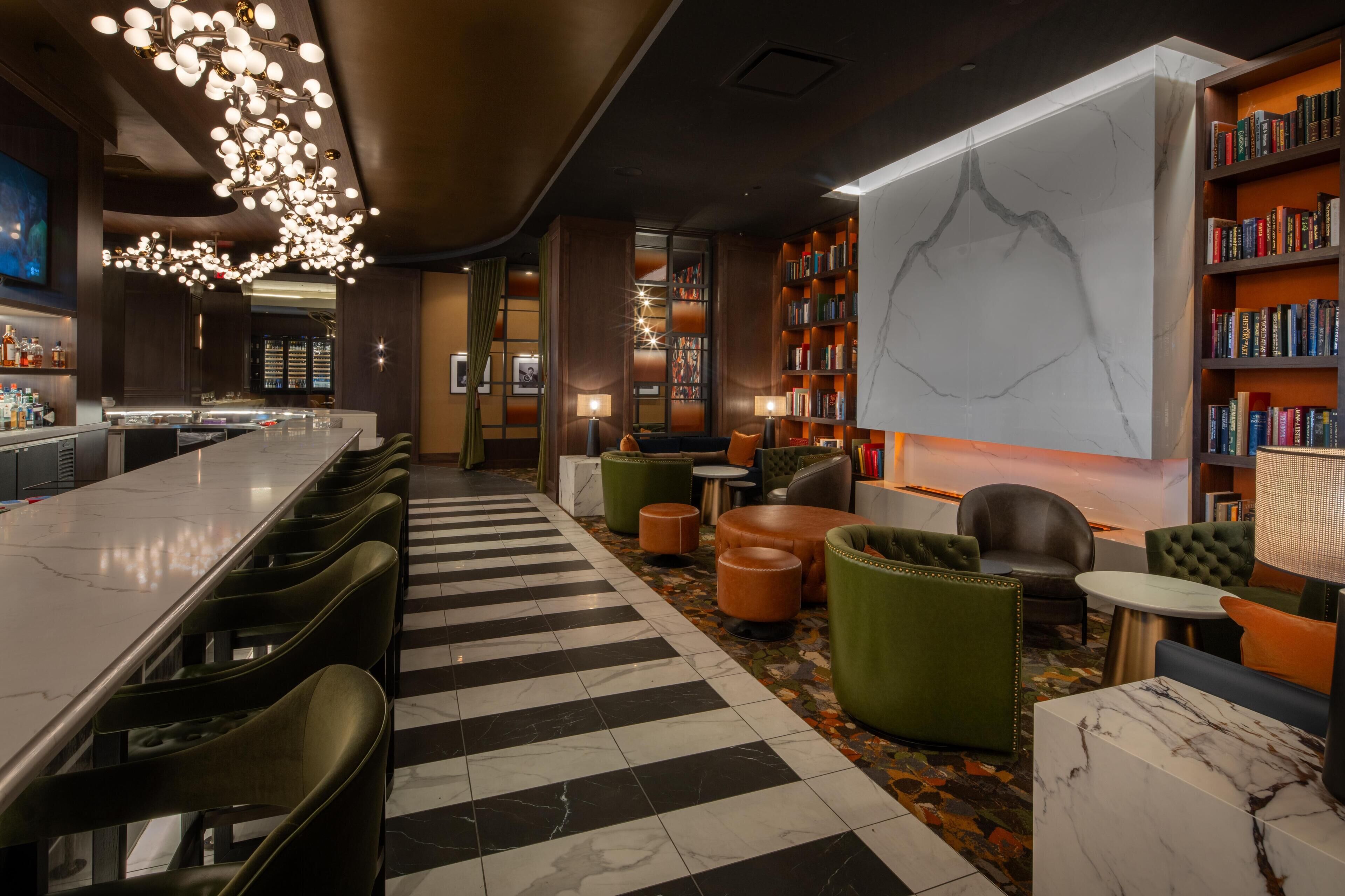 A modern bar area with a long counter, multiple bar stools, a patterned floor, and a large bookshelf filled with books.