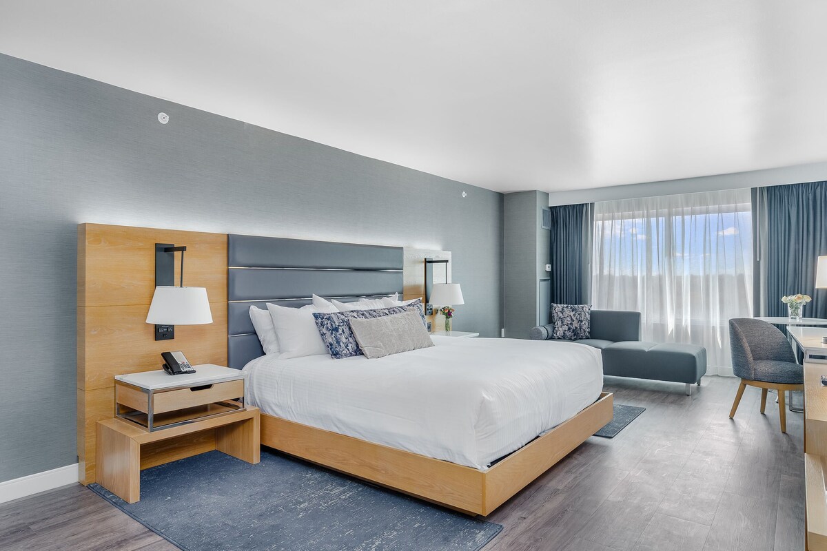 A modern hotel room with a large bed, bedside tables, a desk, and a view of the city.