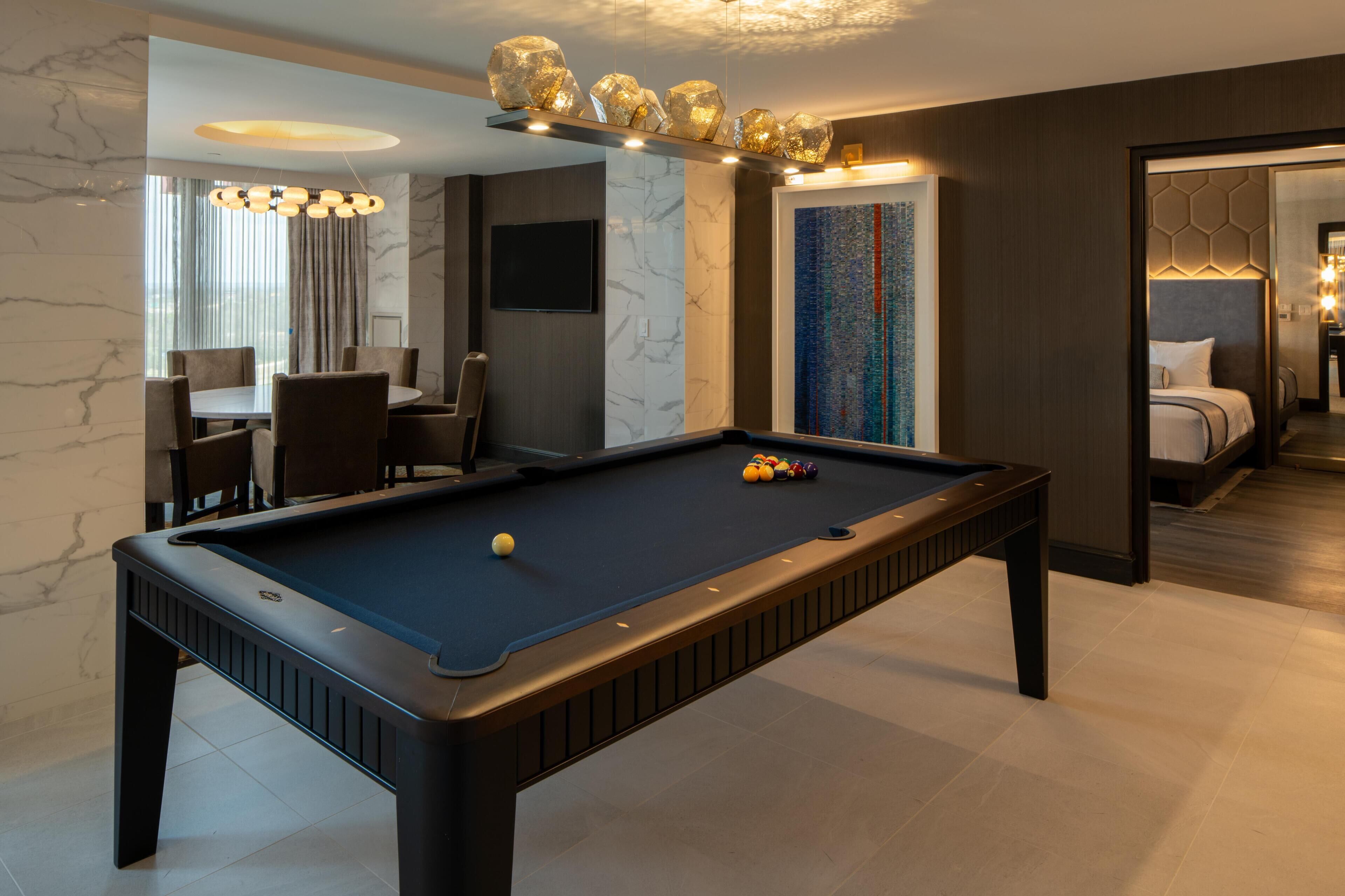 Uma mesa de bilhar com bolas, um espaço de refeições com cadeiras, uma televisão e um quadro.