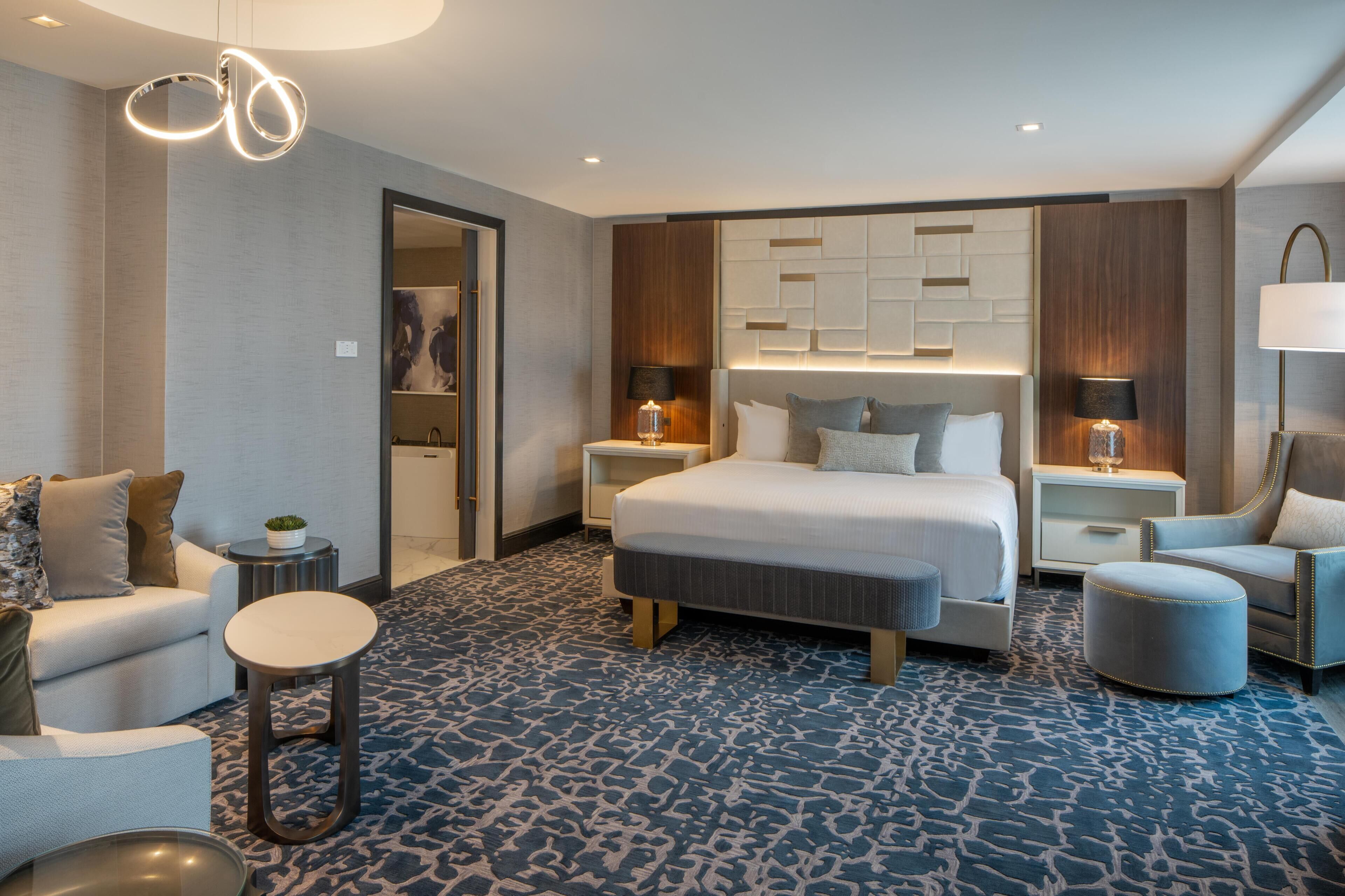 A modern hotel room with a large bed, two bedside tables, a sofa, a round coffee table, and a patterned carpet.