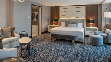 A modern hotel room with a large bed, two bedside tables, a sofa, a round coffee table, and a patterned carpet.