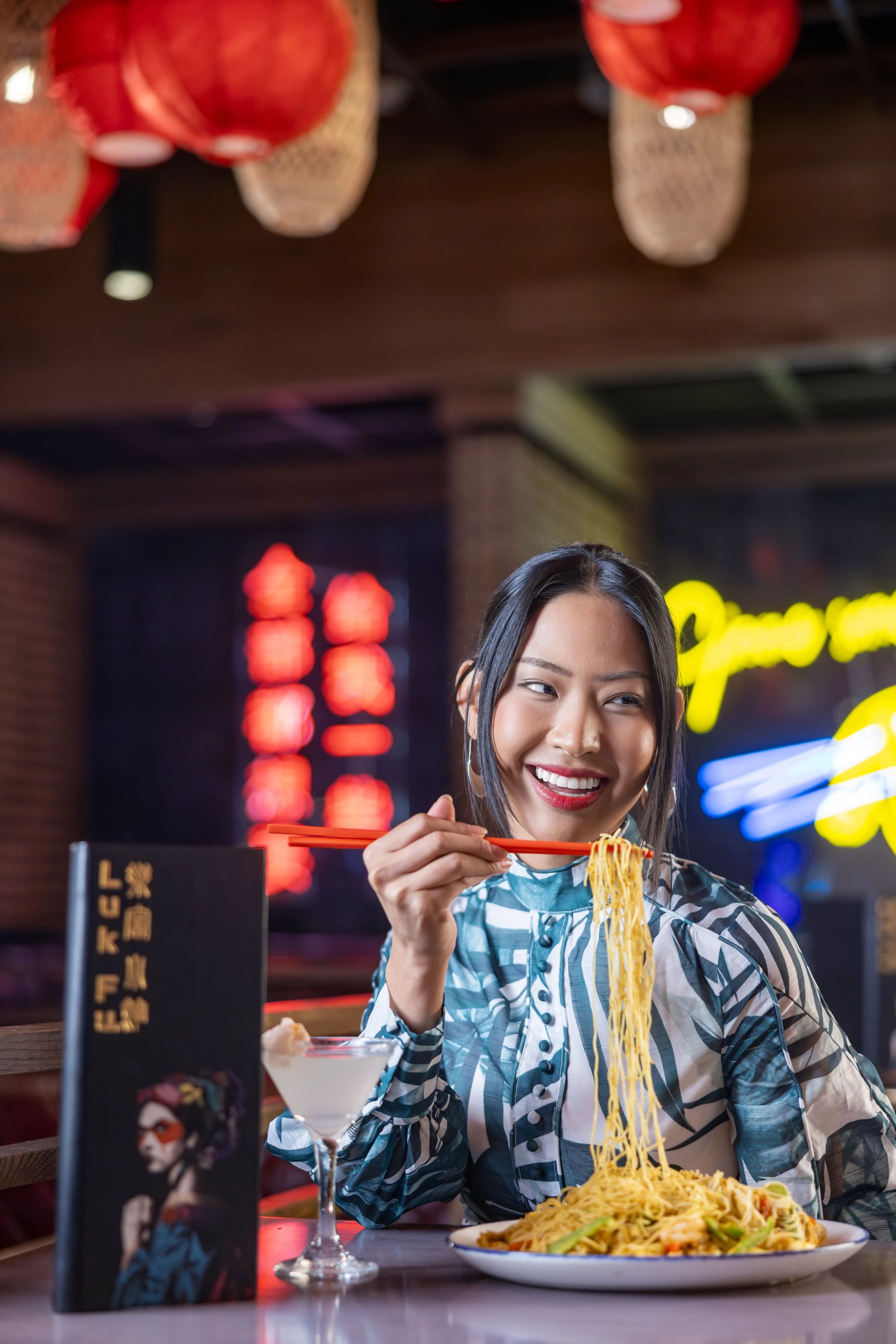 Une personne mangeant des nouilles avec des baguettes, un verre d’une boisson claire et un menu avec des caractères chinois.