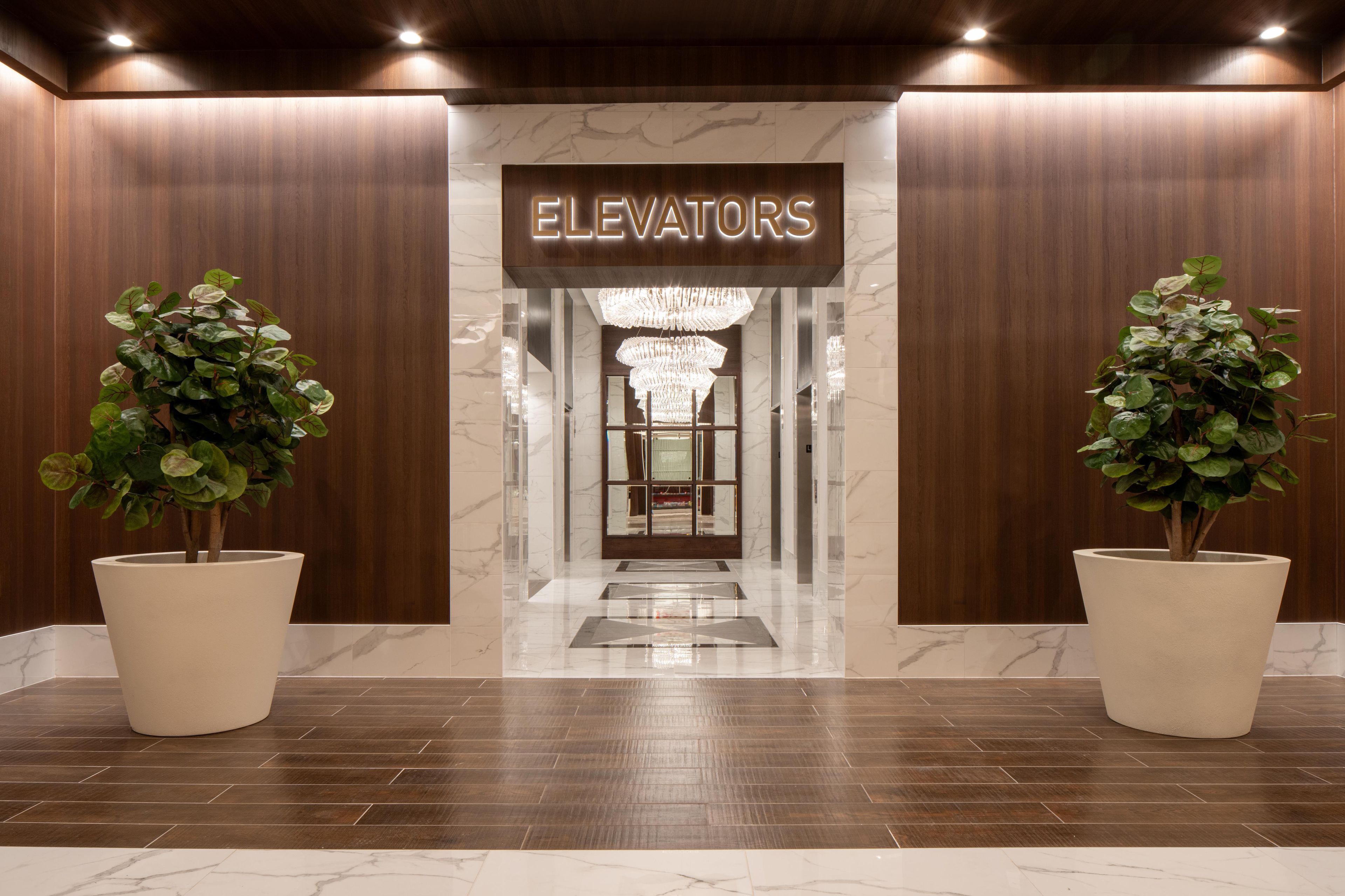 A hotel lobby with a marble floor, two large potted plants, a sign reading 'ELEVATORS', and a chandelier.
