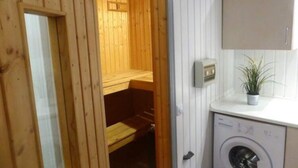 Un sauna en bois avec un banc, un lave-linge et un placard, dans une pièce aux murs blancs.