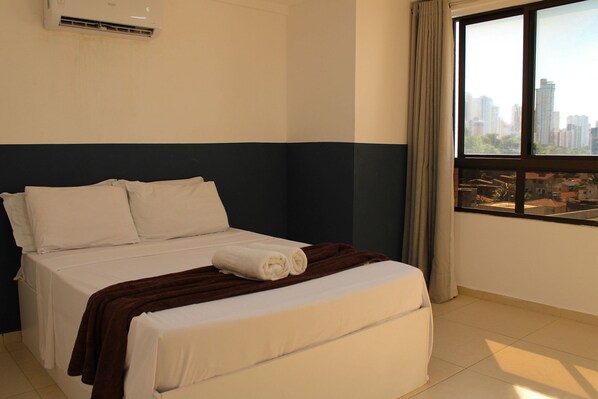 A hotel room with a bed, a window with a city view, and an air conditioning unit. - Flats Villaggio (João Pessoa)