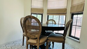 Un comedor con una mesa redonda, cuatro sillas de madera y ventanas amplias con persianas color beige.