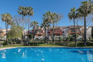 A swimming pool with a clear blue water surface, surrounded by palm trees and a row of multi-story buildings with balconies.