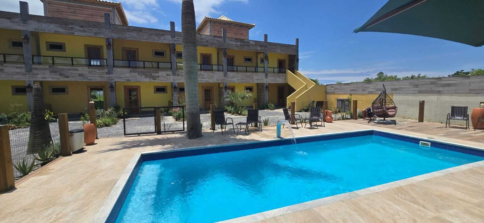 Un área de piscina con una piscina azul cristalina, un edificio amarillo con varios balcones y mobiliario exterior.