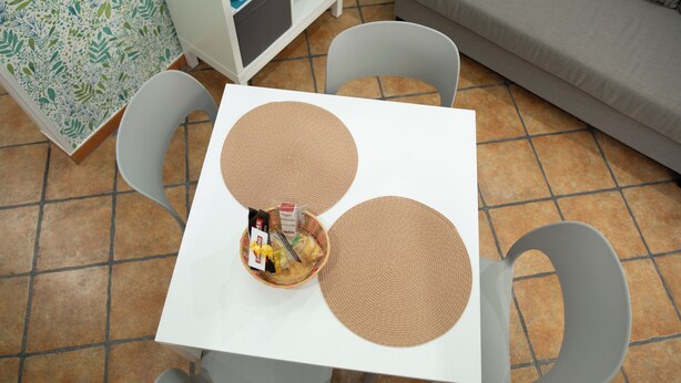 Una mesa blanca con dos individuales tejidos, una cesta con objetos y dos sillas grises.