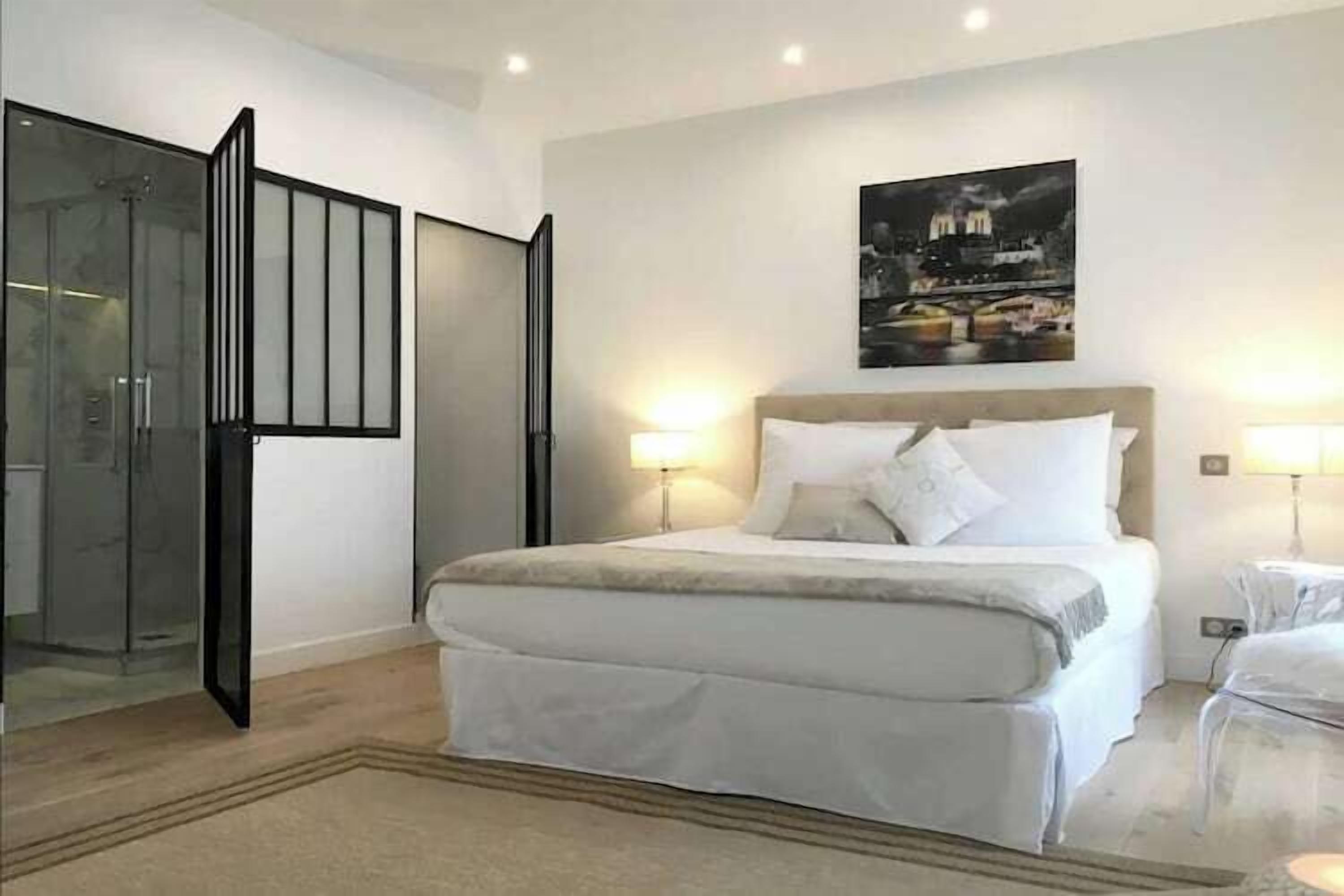 A bedroom with a large bed, two bedside lamps, a nightstand, and a cityscape painting on the wall.