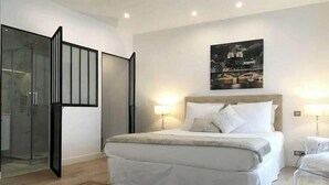 A bedroom with a large bed, two bedside lamps, a nightstand, and a cityscape painting on the wall. - Beautiful apartment - Galeries Lafayette (Paris)