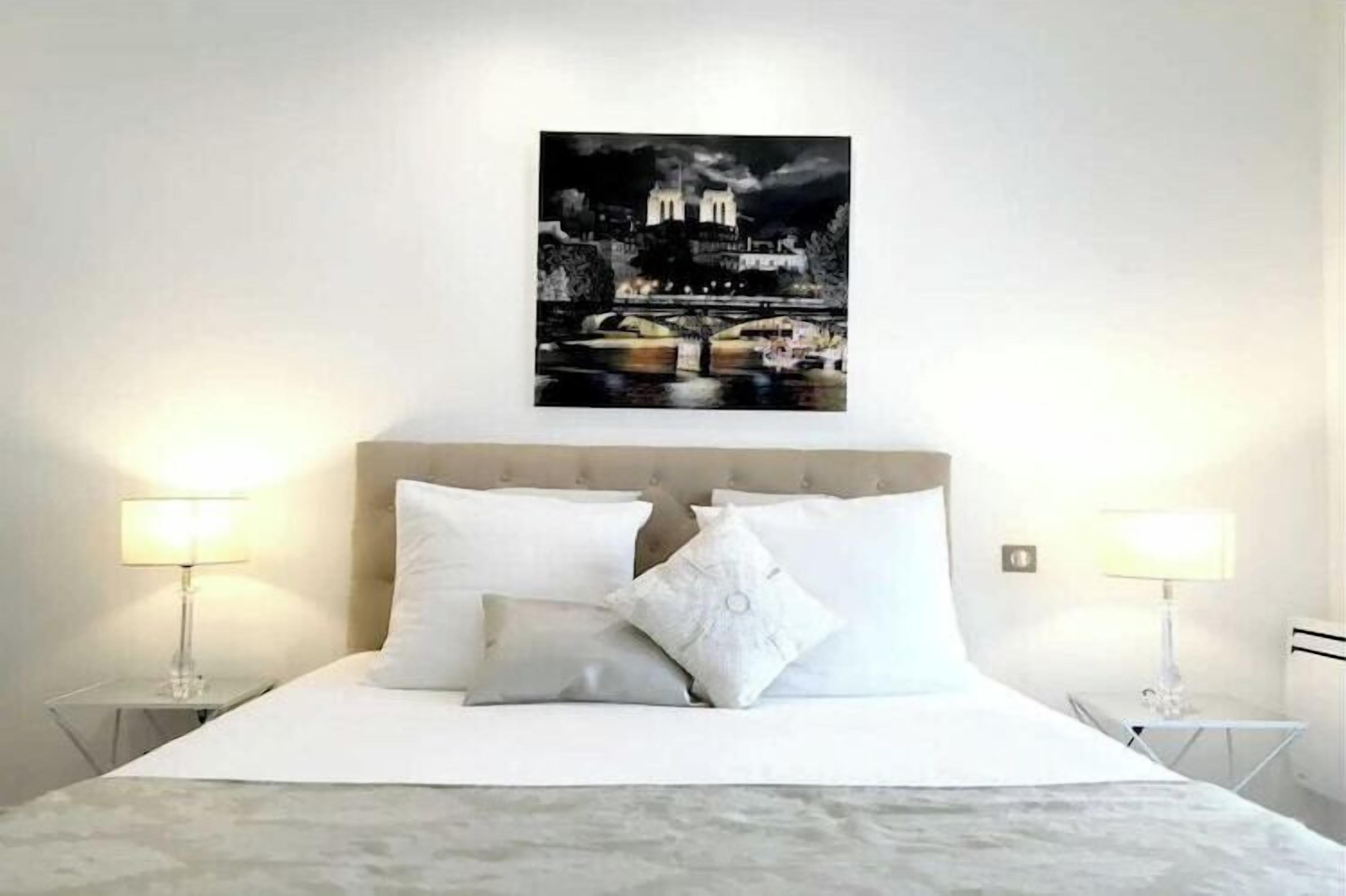 Habitación de hotel con cama, ropa de cama blanca, dos lámparas de noche y un cuadro de un paisaje urbano nocturno en la pared.