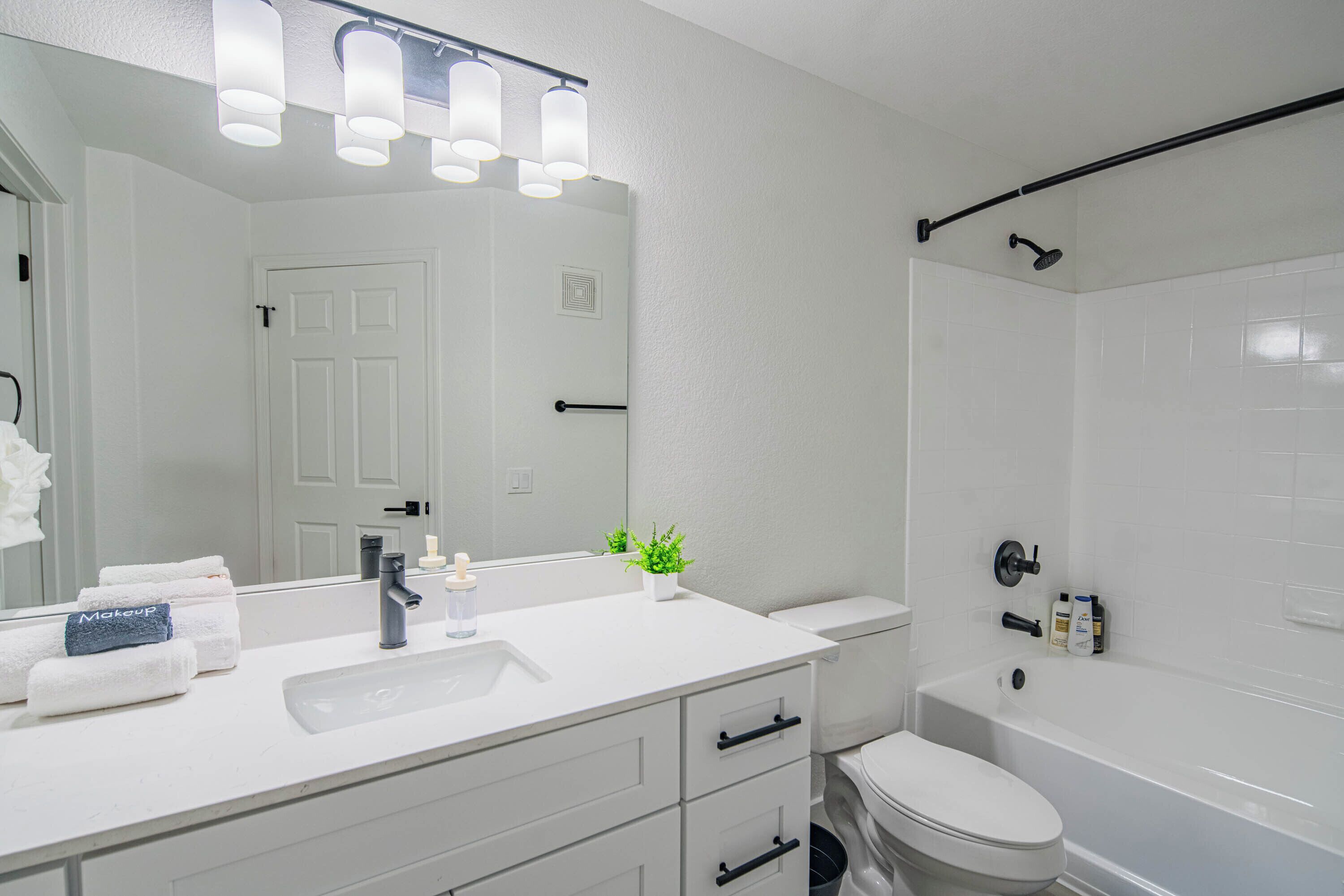 Une salle de bain moderne avec un lavabo, un meuble-lavabo et des toilettes, le tout de couleur blanche. On y trouve également un espace douche avec une tringle à rideau noire et une douchette à main.