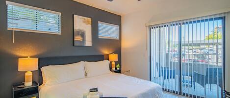 A hotel room with a large bed, two bedside lamps, a ceiling fan, a balcony with a view, and artwork on the walls.