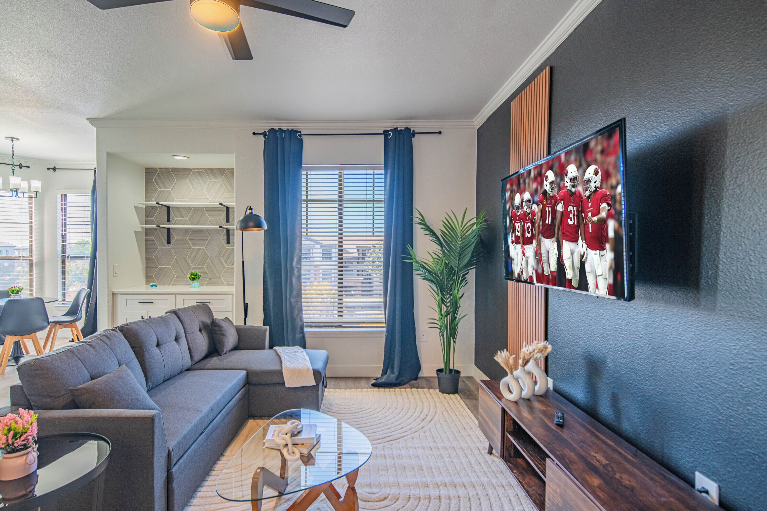 A modern living room with a sectional sofa, a glass coffee table, a flat-screen TV mounted on the wall, a dining area with a kitchen in the background, and a large potted plant.