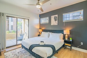 A bedroom with a large bed, two bedside lamps, a ceiling fan, and a view through a sliding glass door to an outdoor area.