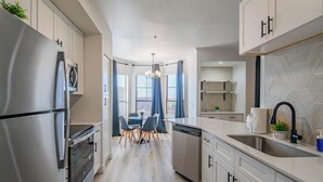 A modern kitchen with stainless steel appliances, a dishwasher, and a sink. There is a dining area with a table and chairs. - ROVE LIVING/KB - Zone (Glendale)
