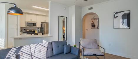 A modern living room with a gray sofa, a black coffee table, and a white armchair. There is a kitchen in the background with white cabinets and a microwave.