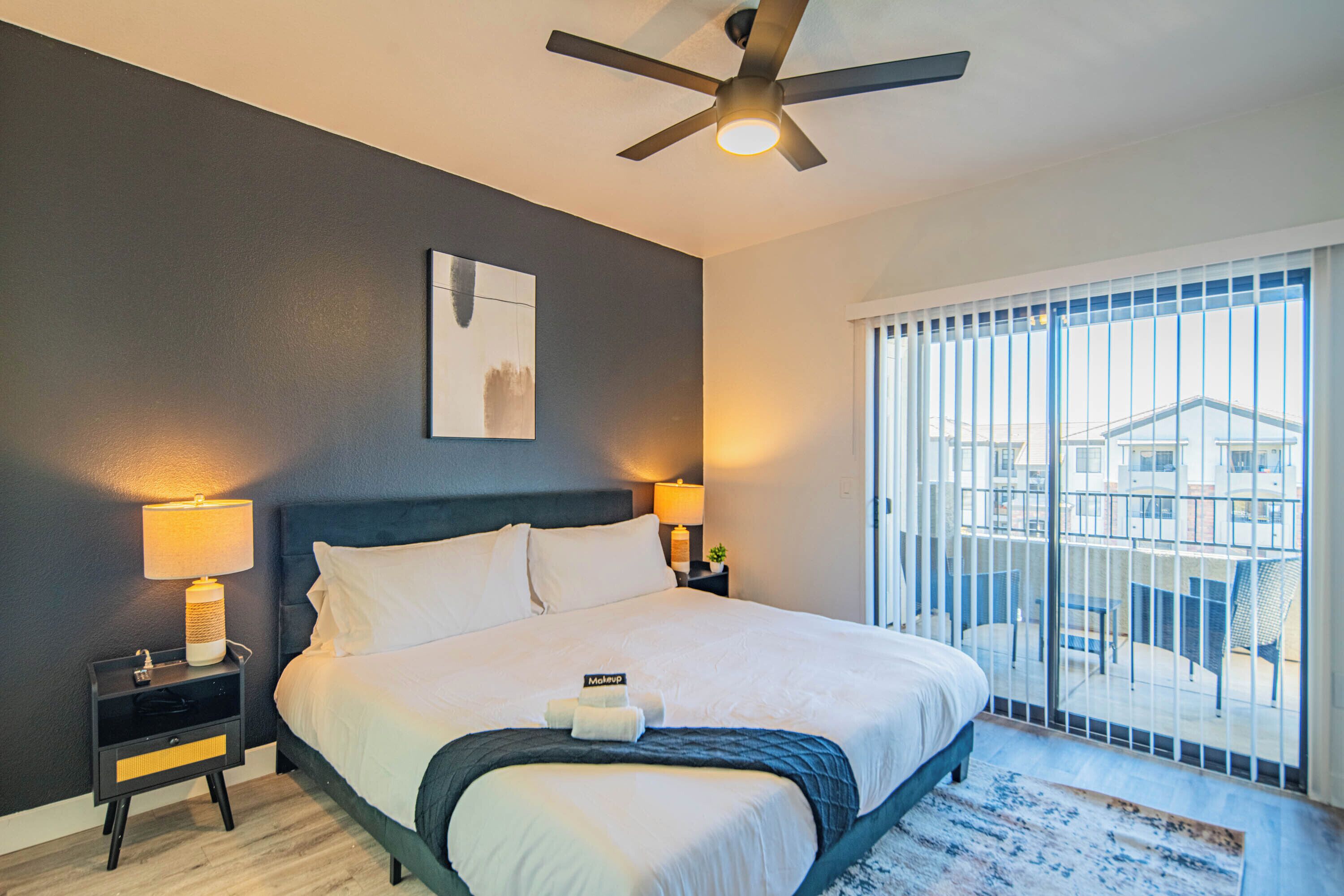 A hotel room with a bed, bedside lamps, a ceiling fan, and a view through a window with blinds.