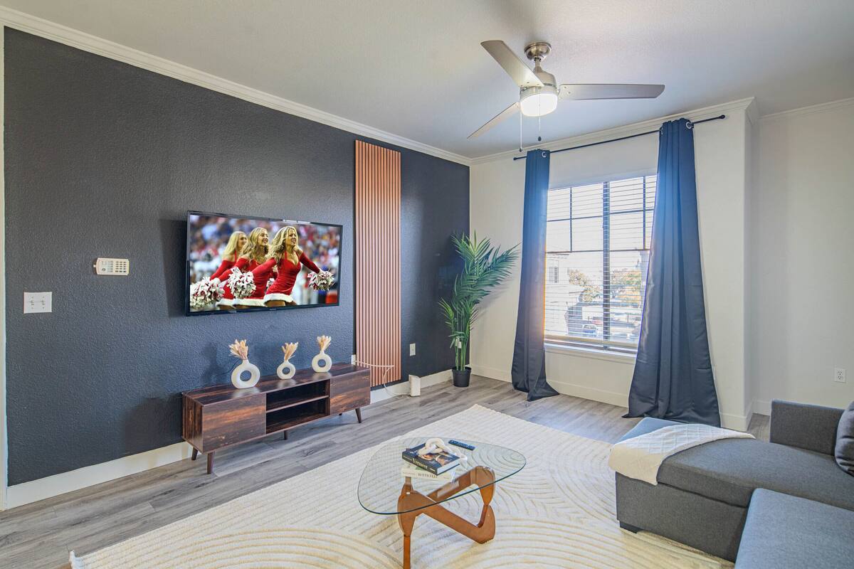 Een moderne woonkamer met een flatscreen televisie, een plafondventilator, een bank, een salontafel en een groot raam met gordijnen.