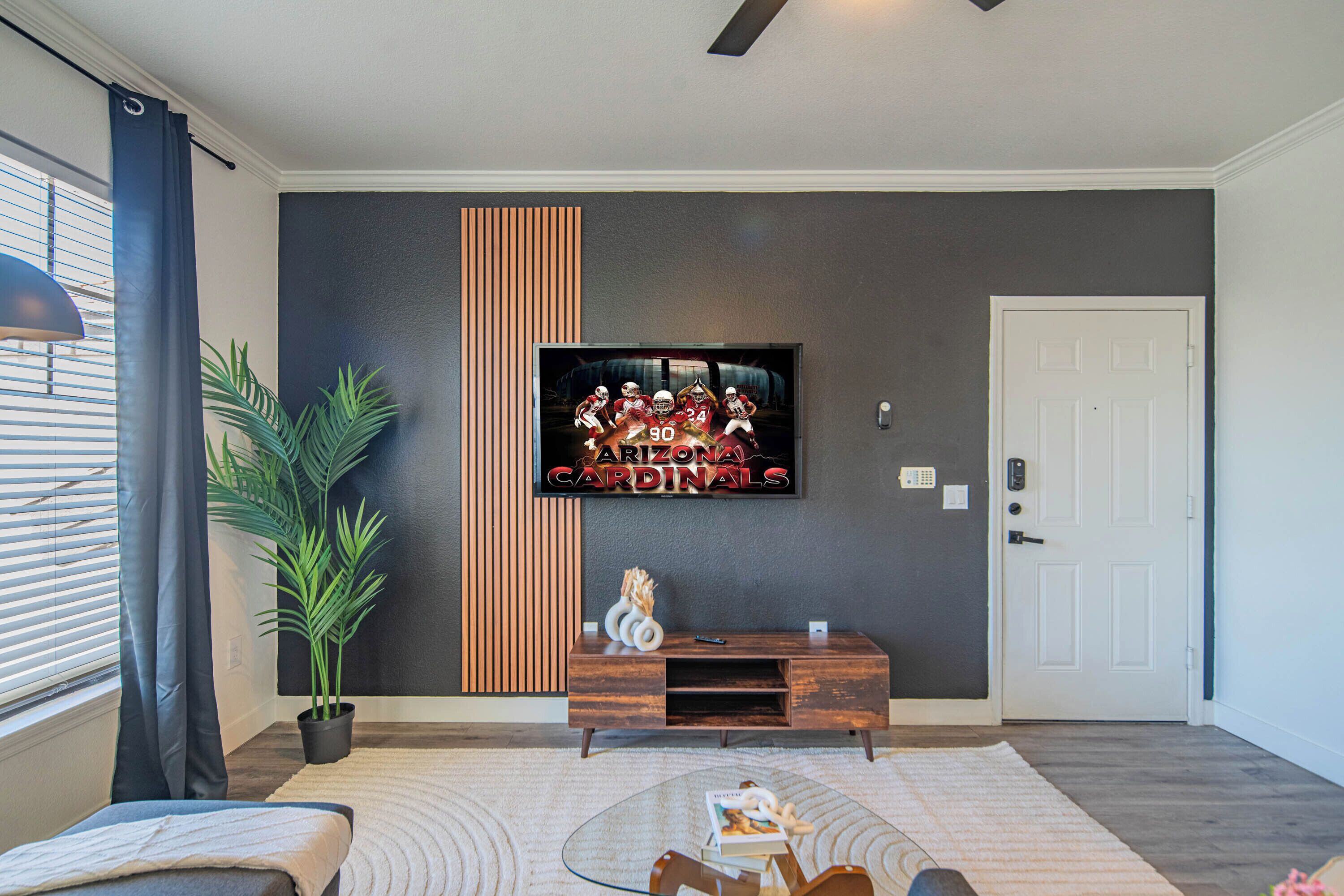 A modern living room with a flat-screen TV mounted on the wall, a wooden entertainment unit, a glass coffee table, and a potted plant.