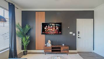 A modern living room with a flat-screen TV mounted on the wall, a wooden entertainment unit, a glass coffee table, and a potted plant.