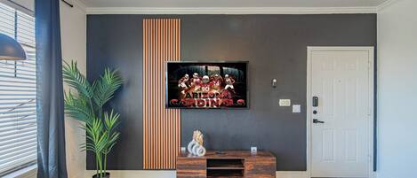 A modern living room with a flat-screen TV mounted on the wall, a wooden entertainment unit, a glass coffee table, and a potted plant.