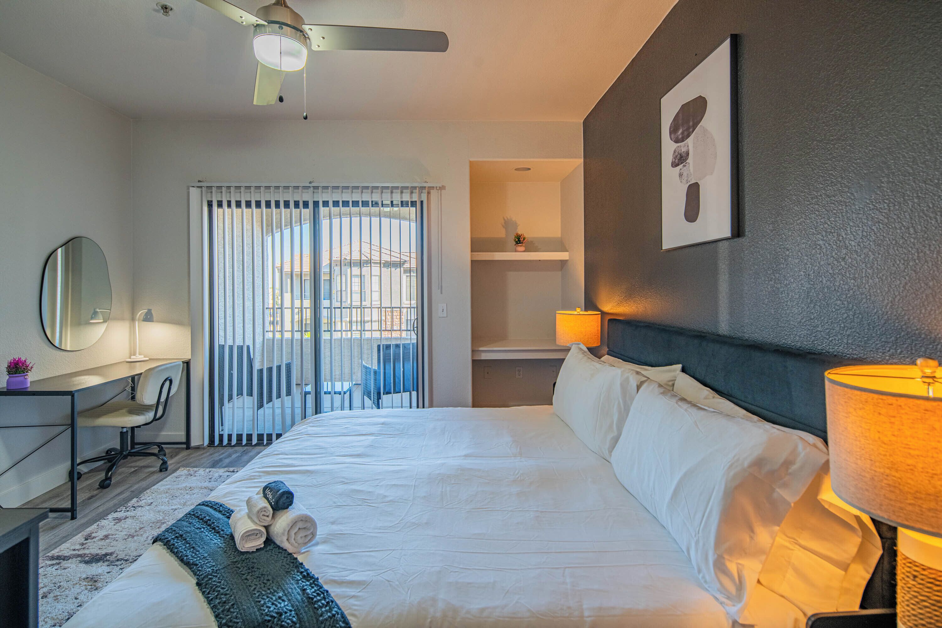 A hotel room with a bed, desk, chair, and a balcony with a view.