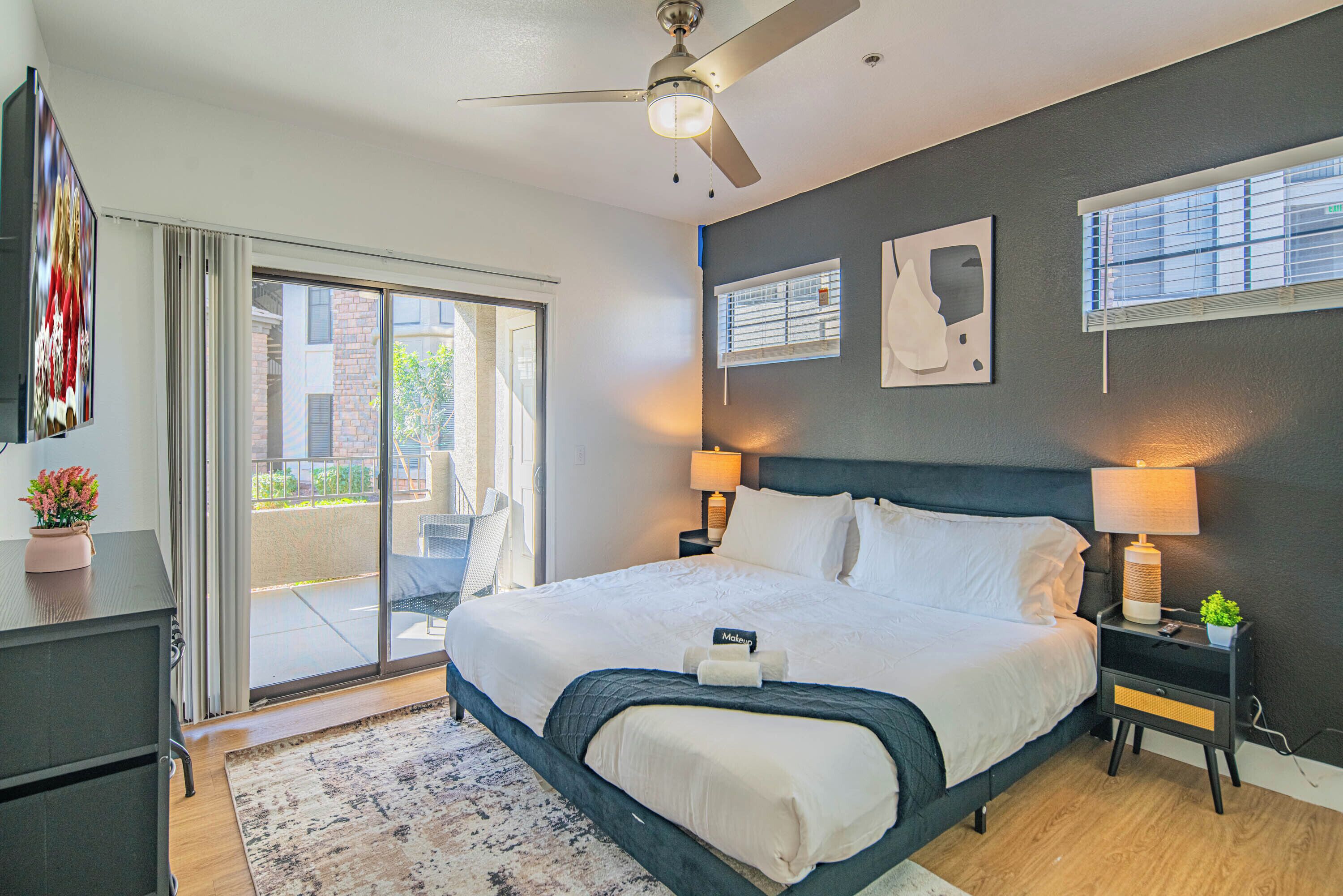 A modern hotel room with a large bed, a ceiling fan, a sliding glass door to the balcony, and a small table with a lamp and a plant.