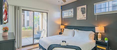 A modern hotel room with a large bed, a ceiling fan, a sliding glass door to the balcony, and a small table with a lamp and a plant.