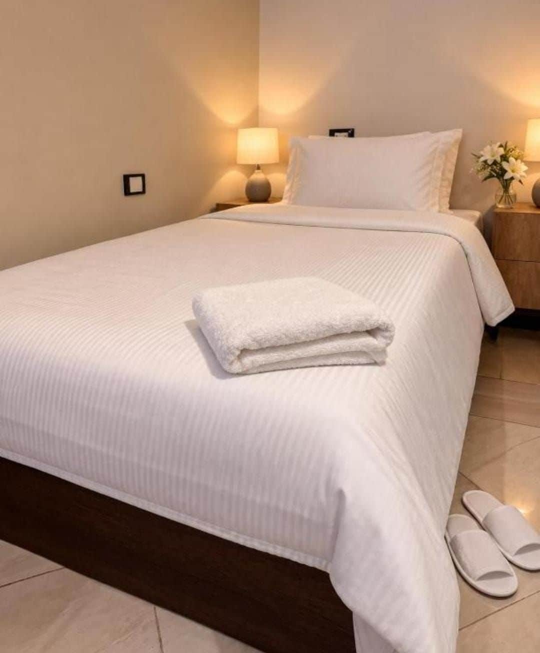 A neatly made bed with a folded white towel, a wooden headboard, and a nightstand with a lamp and a vase of flowers.