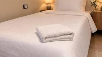 A neatly made bed with a folded white towel, a wooden headboard, and a nightstand with a lamp and a vase of flowers.