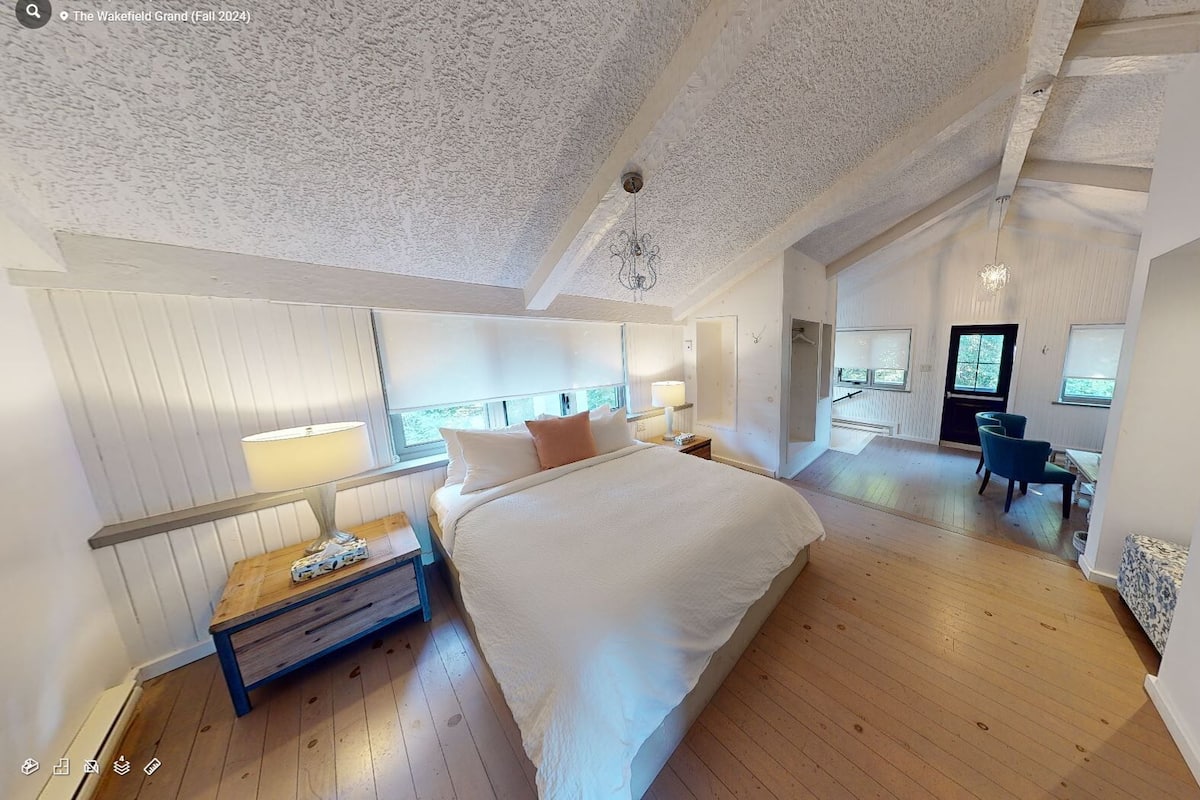 A spacious bedroom with a large bed, a desk, and a chair, featuring a sloped ceiling and large windows.