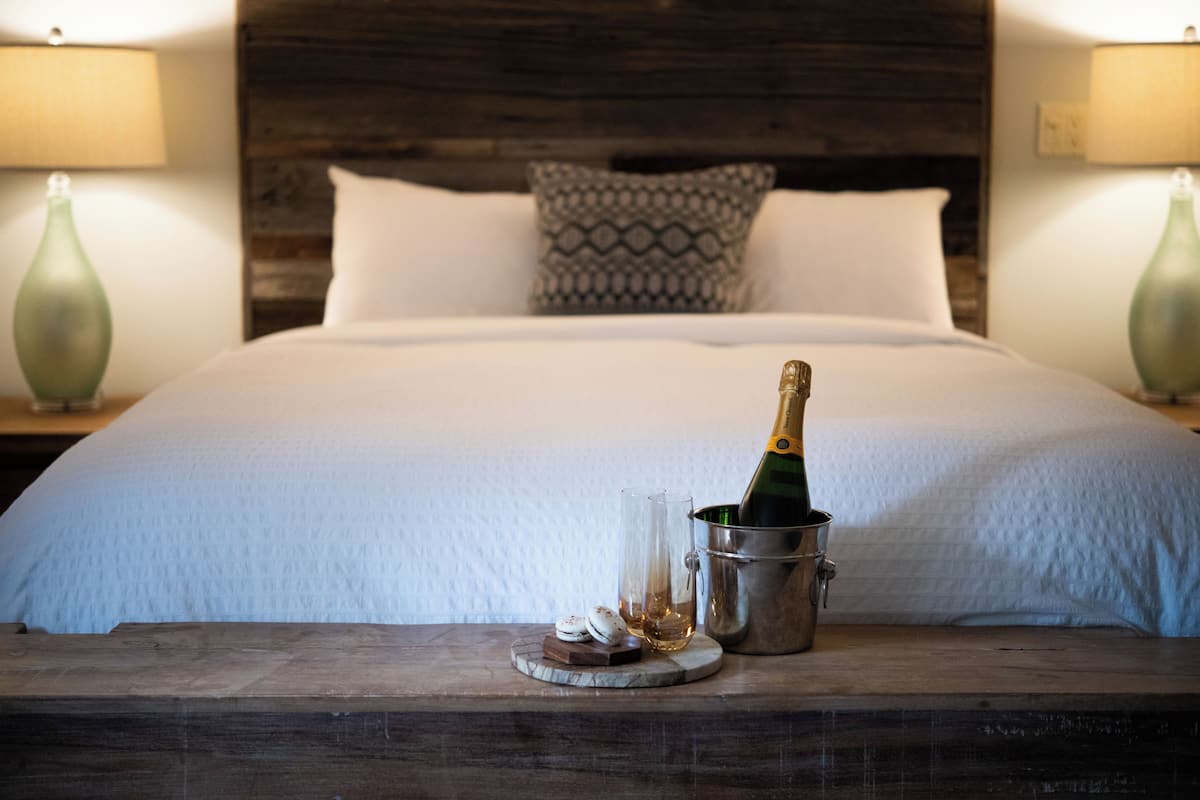 A neatly made bed with a wooden headboard, a bottle of champagne, and a glass on a wooden nightstand.