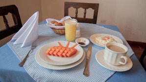 Ein Frühstückstisch mit kariertem Tischläufer, einer Speisenplatte, einem Glas Saft, einer Tasse Kaffee und einem Korb mit Croissants.