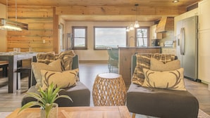 Un interior de cabaña acogedor con zona de comedor, dos sofás con cojines estampados, una mesa redonda de fibras naturales y una cocina con refrigerador.