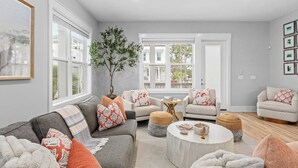 Ein modernes Wohnzimmer mit einem grauen Sofa, zwei Sesseln, einem Couchtisch und einem großen Fenster mit Jalousien.